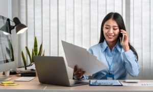 Mulher sorridente vestindo uma camisa azul e usando um crachá, sentada à mesa de trabalho. Ela está ao telefone e segurando um documento. Na mesa, há um laptop, uma luminária, alguns papéis e uma planta decorativa ao fundo