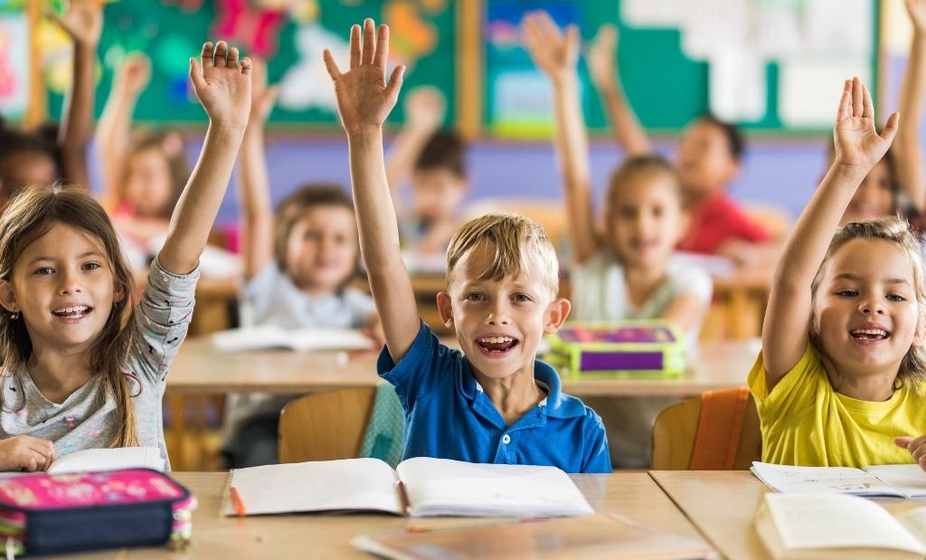 crianças em sala de aula participando ativamente, levantando as mãos para responder, em um ambiente escolar alegre e interativo, com livros abertos na mesa, destacando a valorização da educação infantil e ensino fundamental.