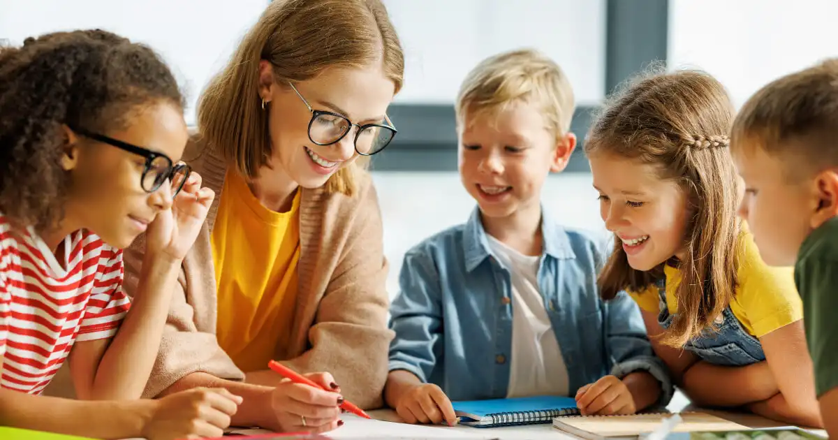 Imagem de uma professora sorridente interagindo com um grupo de crianças na sala de aula durante uma atividade escolar divertida e educativa.