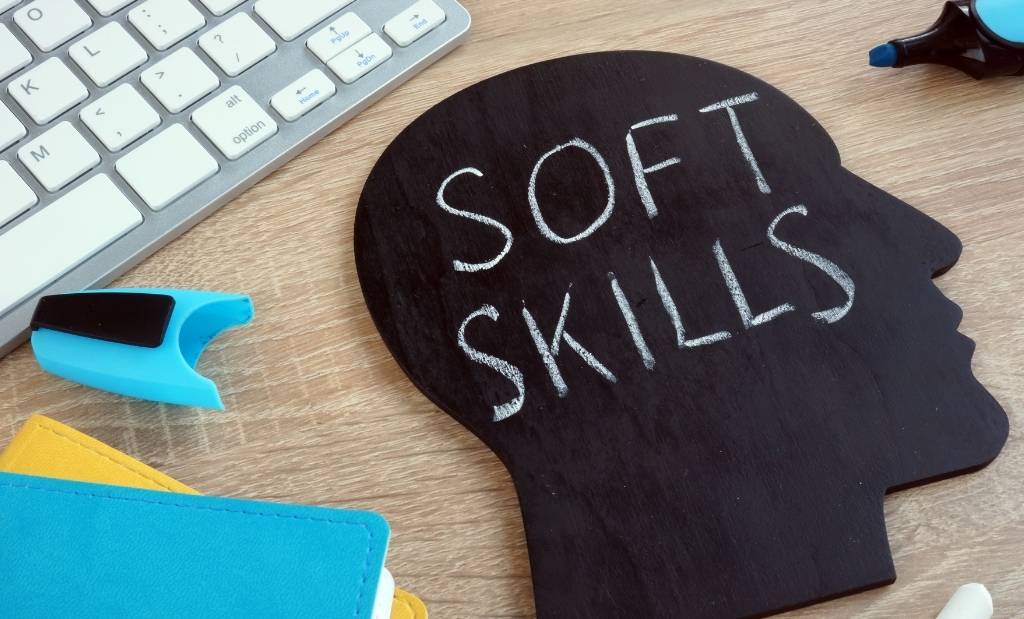placa preta com a inscrição "soft skills" sobre uma mesa de trabalho com teclado e materiais escolares.