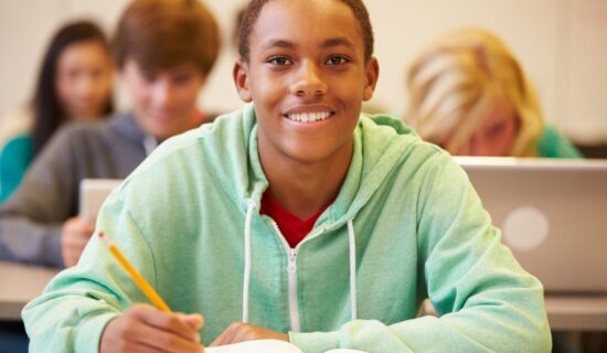 estudante sorridente escrevendo em sala de aula com colegas ao fundo.