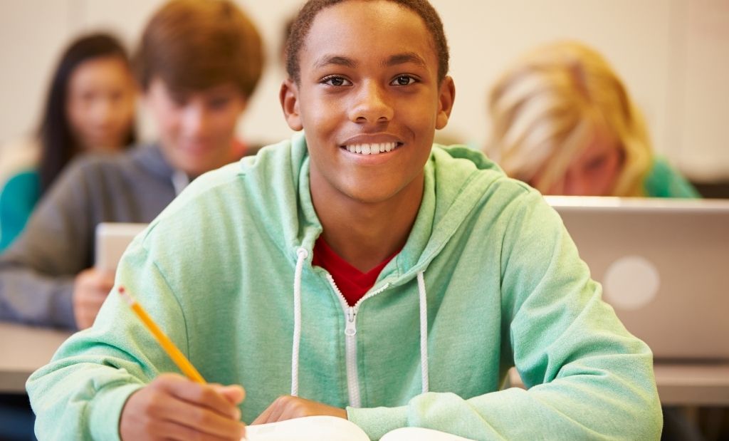 estudante sorridente escrevendo em sala de aula com colegas ao fundo.