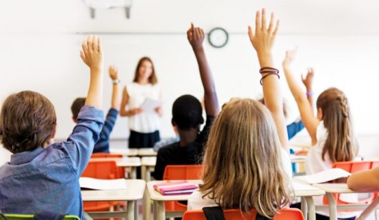 Uma sala de aula cheia de crianças, vista por trás, com várias mãos levantadas em sinal de participação ou para fazer perguntas. No fundo da sala, de forma ligeiramente desfocada, uma professora está em pé na frente de uma lousa branca, segurando papéis e olhando para os alunos.