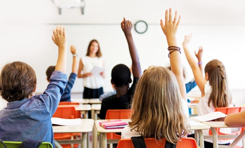 Uma sala de aula cheia de crianças, vista por trás, com várias mãos levantadas em sinal de participação ou para fazer perguntas. No fundo da sala, de forma ligeiramente desfocada, uma professora está em pé na frente de uma lousa branca, segurando papéis e olhando para os alunos.